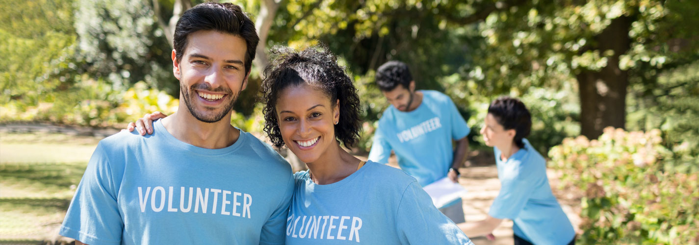 Two smiling volunteers standing in the park, wearing light blue "volunteer" shirts.