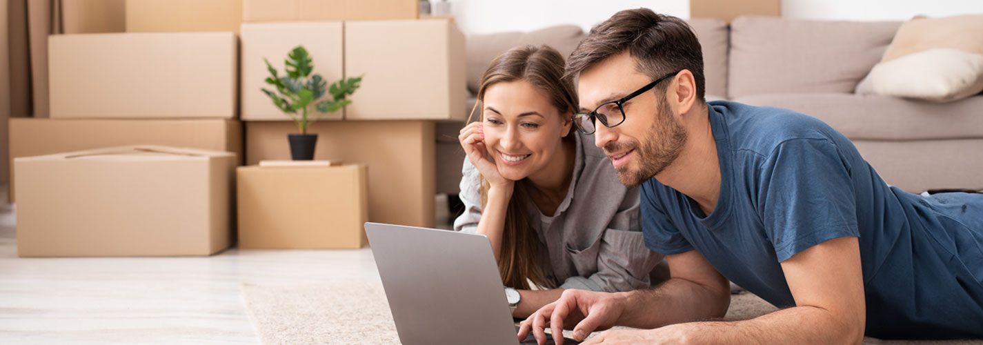 A smiling couple lays on the floor of their living room, surrounded by boxes, in front of their laptop and plans together.