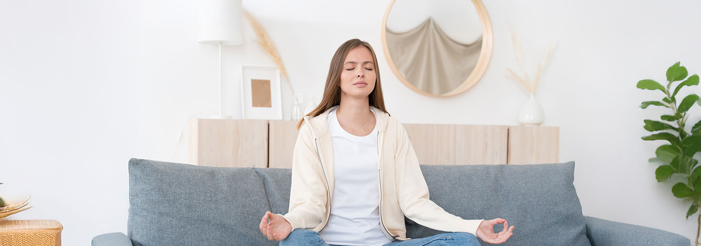 A young woman sitting on her couch in lotus pose and meditating.