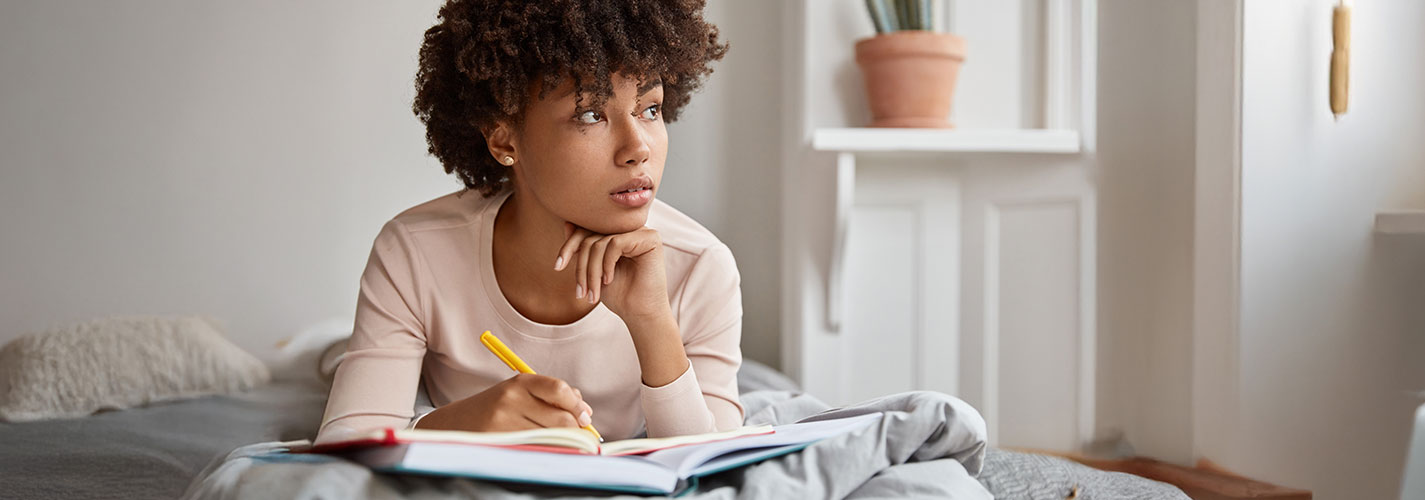 A young woman writing in her journal and looking out to the window reflecting.