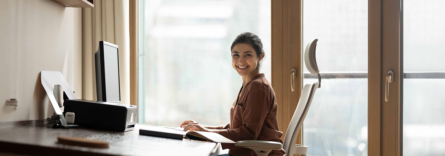 Woman smiling while working at a desk with ergonomic posture in a bright, home office setting.