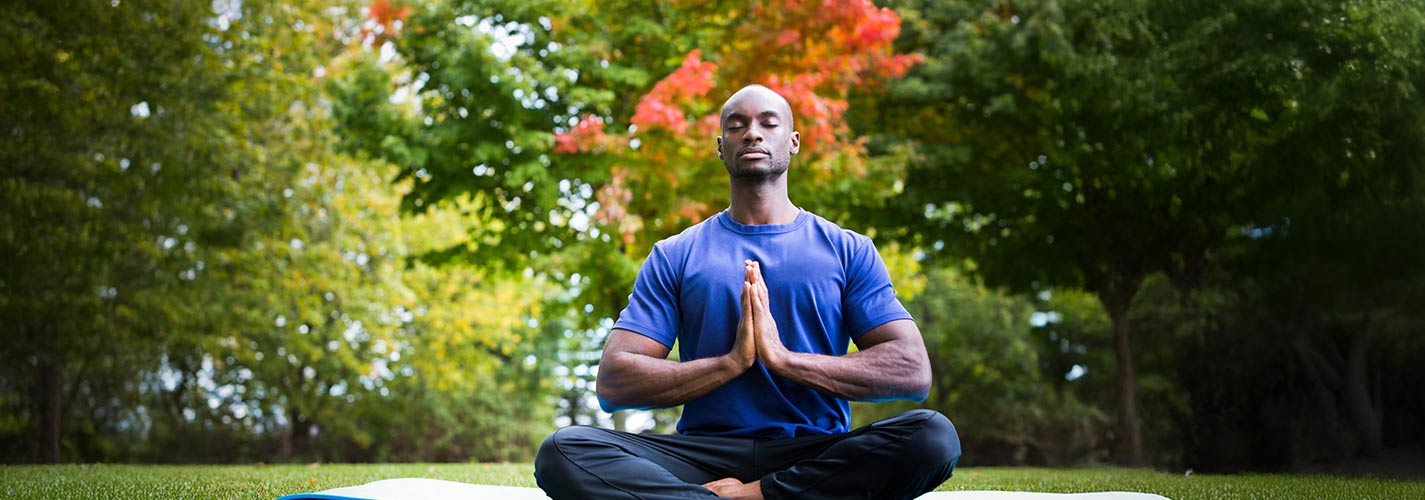 Man meditating cross-legged on a yoga mat in a park, with colorful autumn trees behind him.