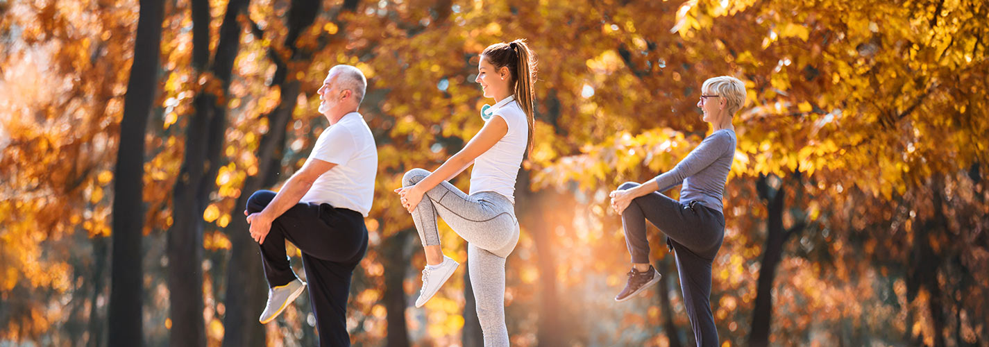 Three people exercising outdoors in a park during autumn, lifting one knee while standing.