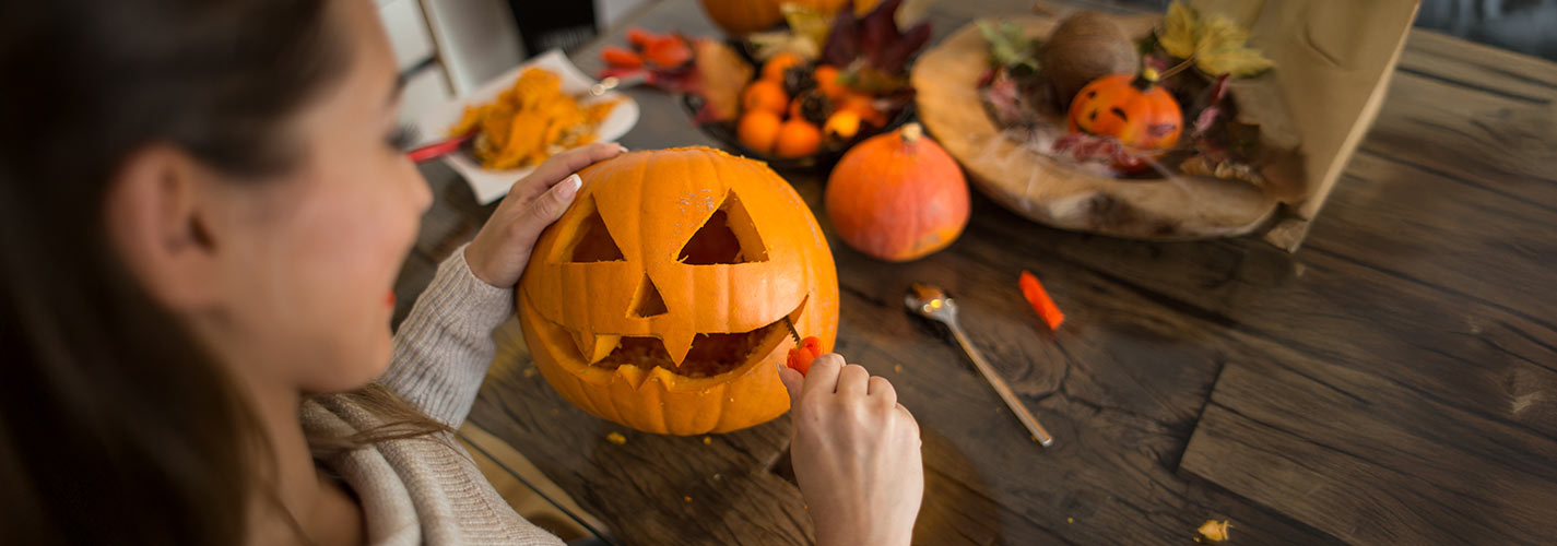 A woman carving a jack-o’-lantern pumpkin at a wooden table decorated with autumn decorations.