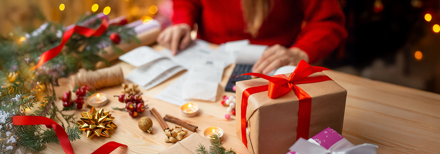 A person sits at a table budgeting with a calculator and receipts, surrounded by holiday decorations and presents.