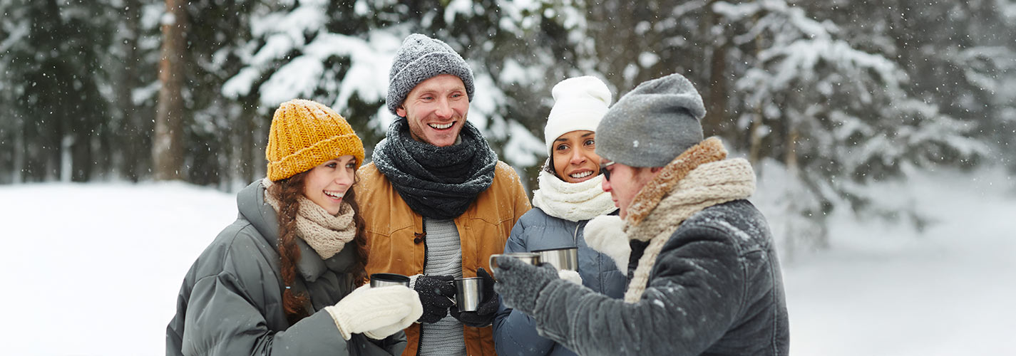 Happy young people standing together drinking hot tea in a thermos in a snowy winter forest.